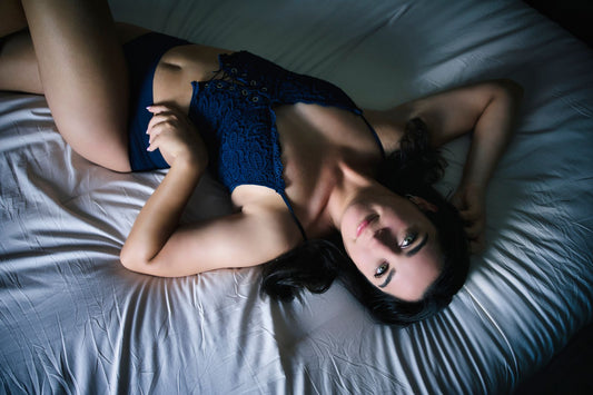 Woman in navy lace lingerie relaxing on white sheets in moody boudoir lighting, a calm self-care moment