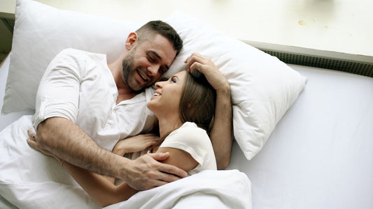 Smiling couple cuddling in bed under white sheets — relaxed, playful intimacy