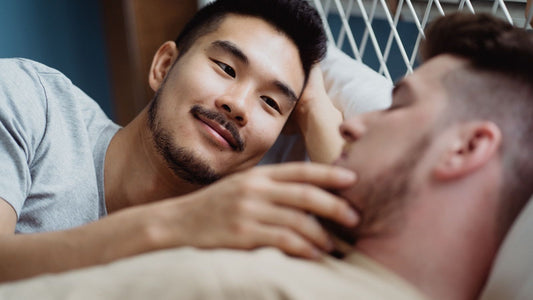 Male couple lying close in bed, one gently touching the other's face — tender gay intimacy