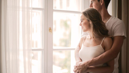 Partner hugging a woman from behind by a sunlit window — tender aftercare closeness