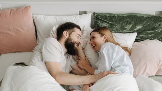 Couple laughing together under the covers in bed — relaxed, shame-free intimacy